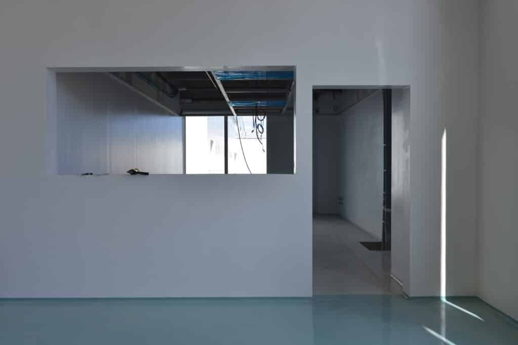 Intérieur en construction: mur blanc avec ouverture, vue sur cables et extérieur. Porte vers pièce sombre. Sol bleu-vert brillant. Lumière rasante.