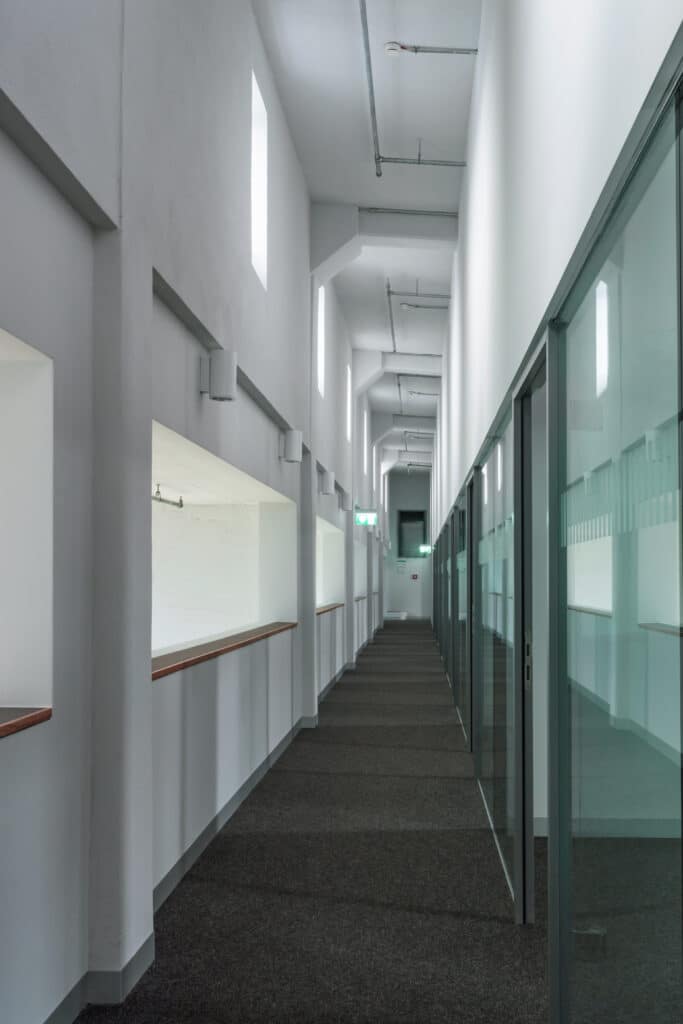 Couloir moderne et lumineux avec murs vitrés, ouvertures, moquette, tuyaux apparents et panneaux sortie.