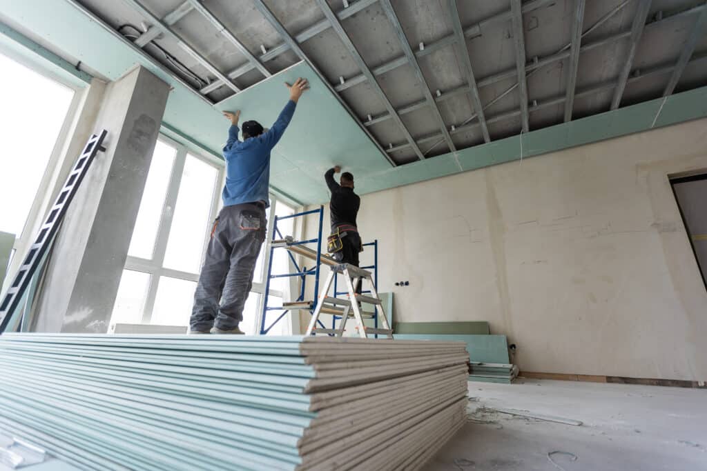 Deux ouvriers installent du placoplatre sur un plafond avec structure métallique sur un chantier de rénovation. Pile de plaques.