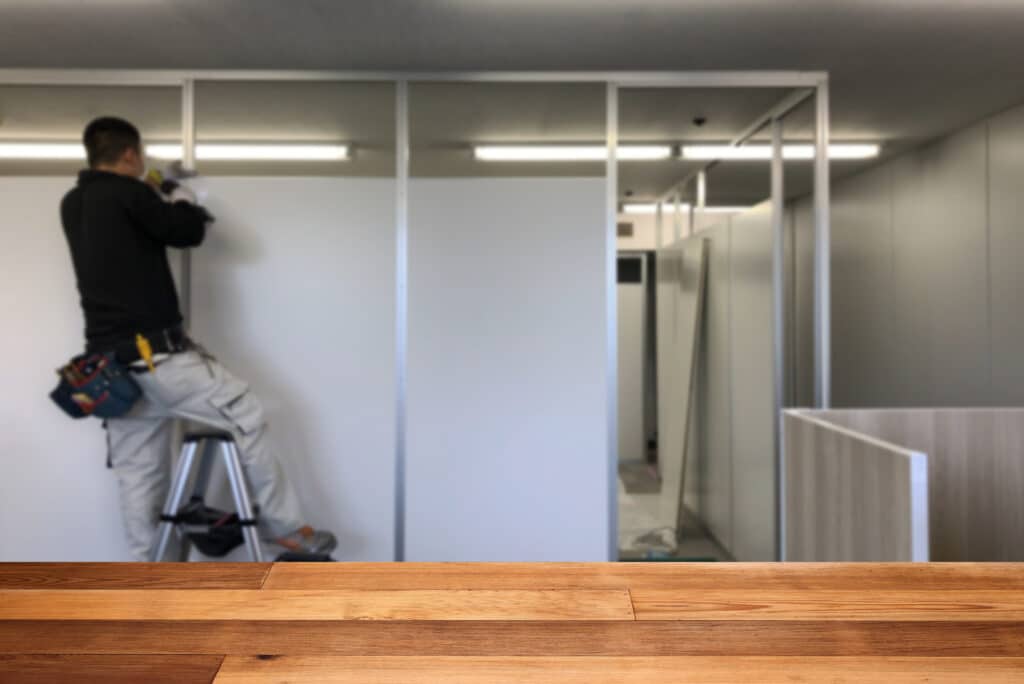 Homme sur escabeau installant une cloison de bureau avec une perceuse. Rénovation d'espace de travail, sol en bois au premier plan.