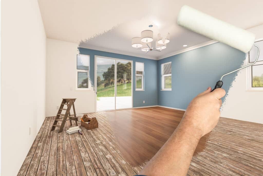 Rénovation d'une chambre : mur en partie peint en bleu par une main au rouleau, sol bois transformé, échelle et outils.
