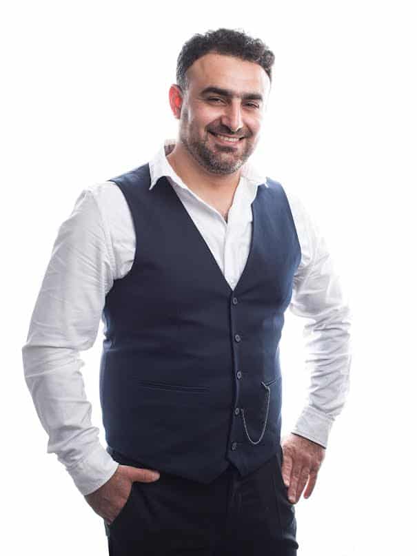 Homme barbu souriant en gilet bleu marine et chemise blanche, main dans la poche. Look élégant et amical.